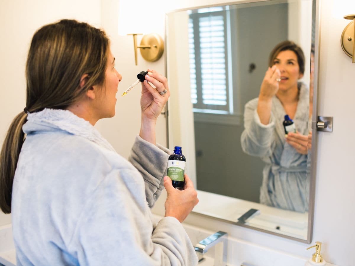 A woman taking CBD oil under her tongue in front of a mirror.