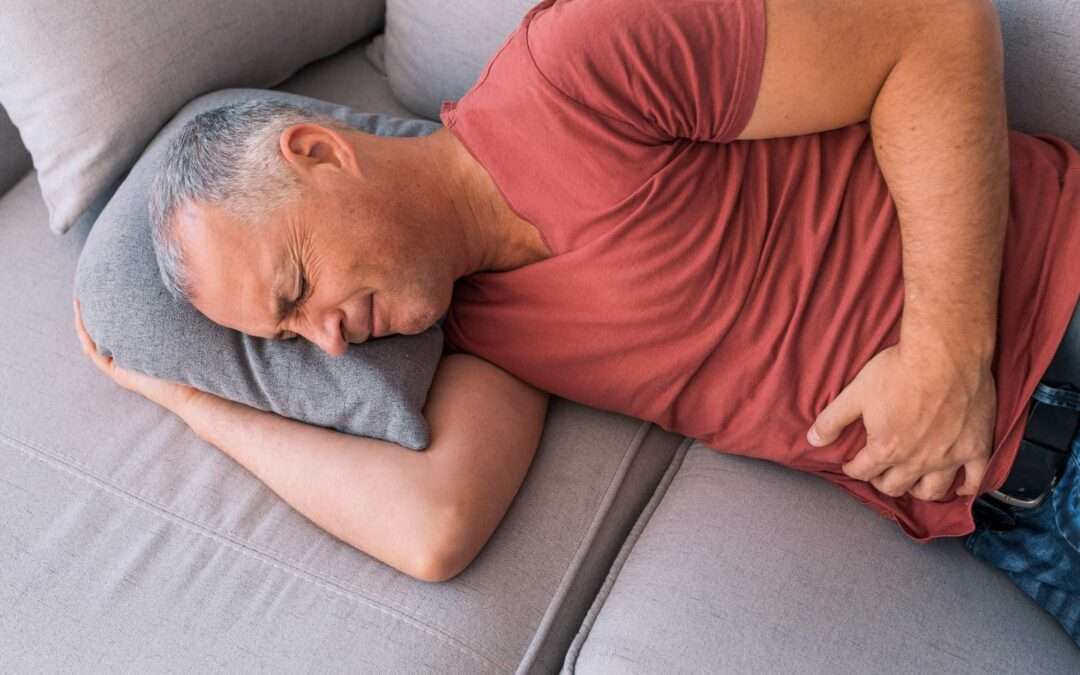 Man lying on a couch, clutching his lower abdomen in discomfort, depicting symptoms of Chronic Prostatitis/Chronic Pelvic Pain Syndrome.